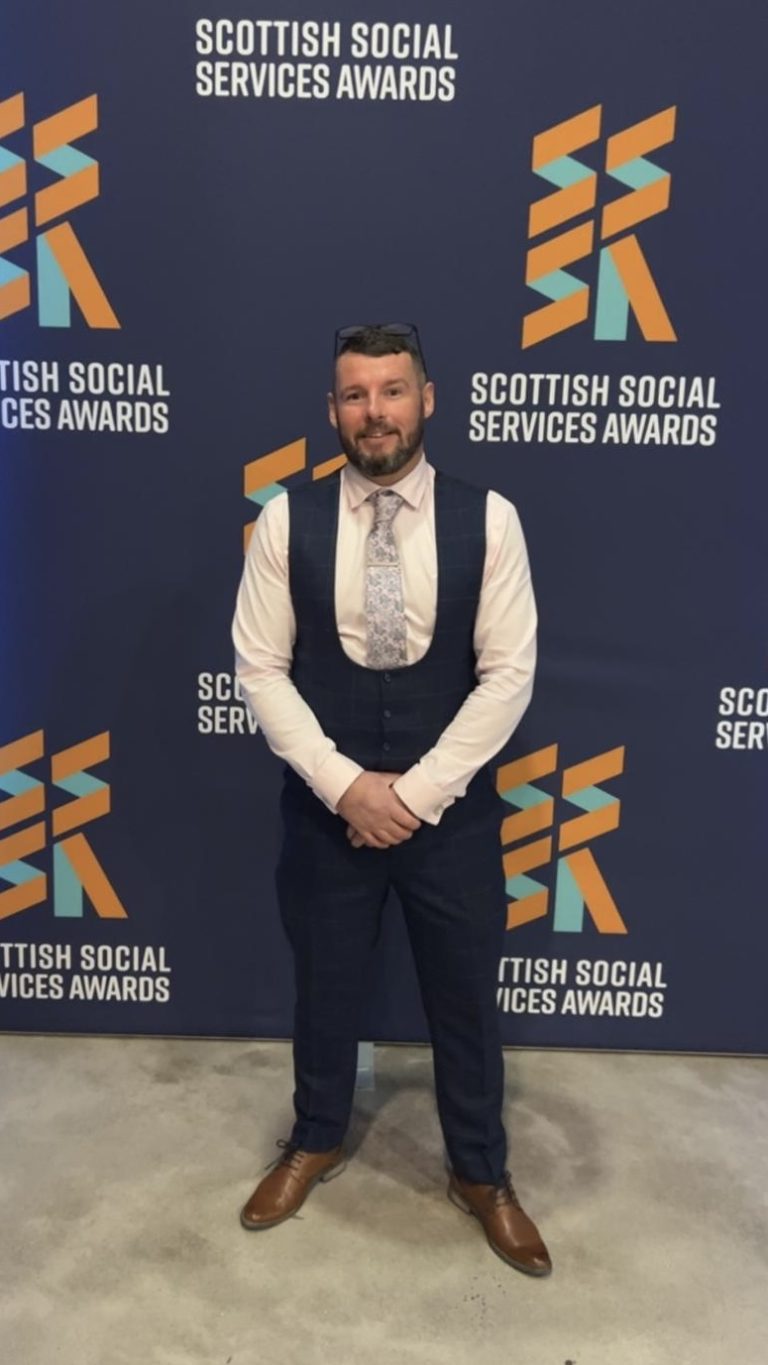 Photo of Scott in a suit and tie, standing in front of a promotional backdrop.