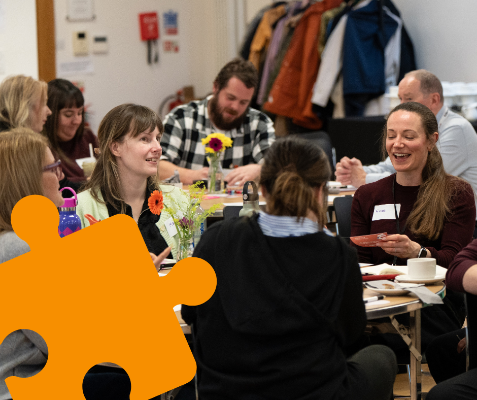 Event participants sitting around tables chatting and writing. A graphic of a small orange jigsaw piece is in the bottom left hand corner of the photo.