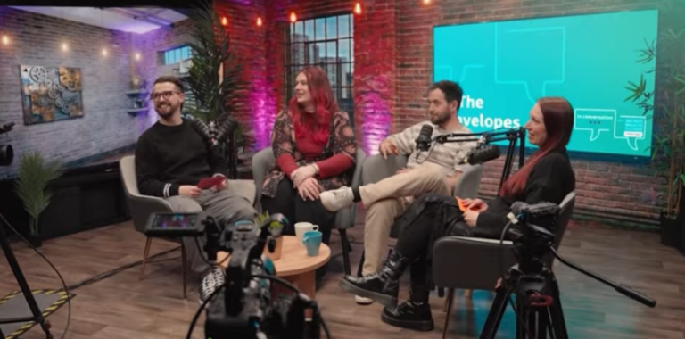 Four people sit in chairs in a studio setting with microphones and cameras, engaged in what appears to be a recording session.