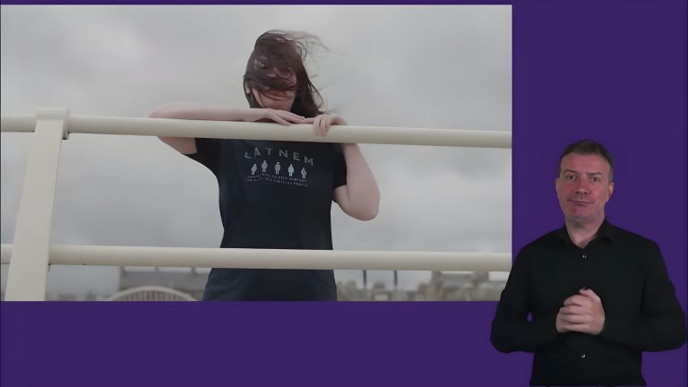 A video still shows a person outdoors leaning on a white railing with their hair blowing in the wind, wearing a black T-shirt with the word "LATNEM" printed above five small human-shaped icons. To the right, a person in a black shirt stands against a solid purple background, performing sign language.